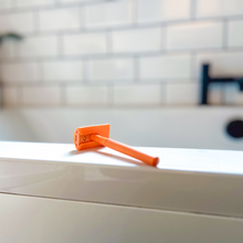 Load image into Gallery viewer, Orange safety razor with matching coloured head protector, lay on the side of a bath - Shoreline Shaving