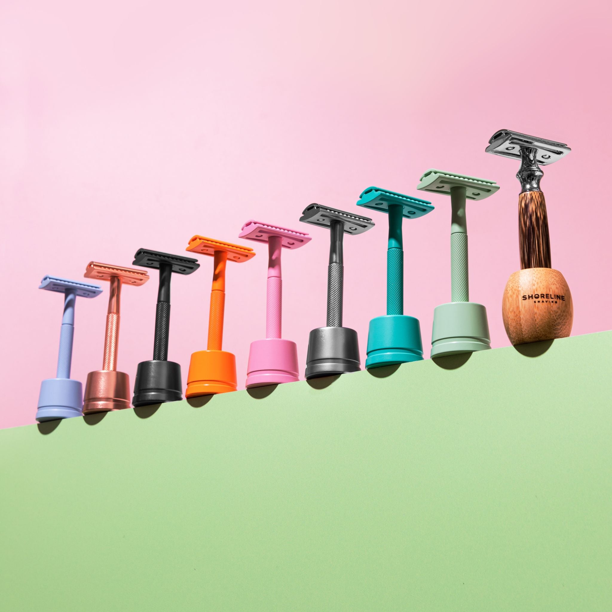 Multi-coloured safety razors in razor stands, lined up on a green base and a pink background - Shoreline Shaving
