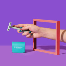 Load image into Gallery viewer, Abstract photo of woman holding a green safety razor through a red square, with a teal blade tin close by - Shoreline Shaving