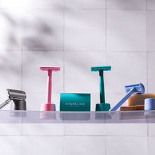 Load image into Gallery viewer, Pale blue safety razor on a bathroom shelf alongside other safety razors in stands, including pink, teal and silver - Shoreline Shaving
